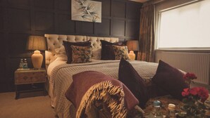 A bedroom with a tufted headboard, a large bed with multiple pillows, a bedside table with a lamp, and a window with curtains. - The Greyhound on the Test (Stockbridge)
