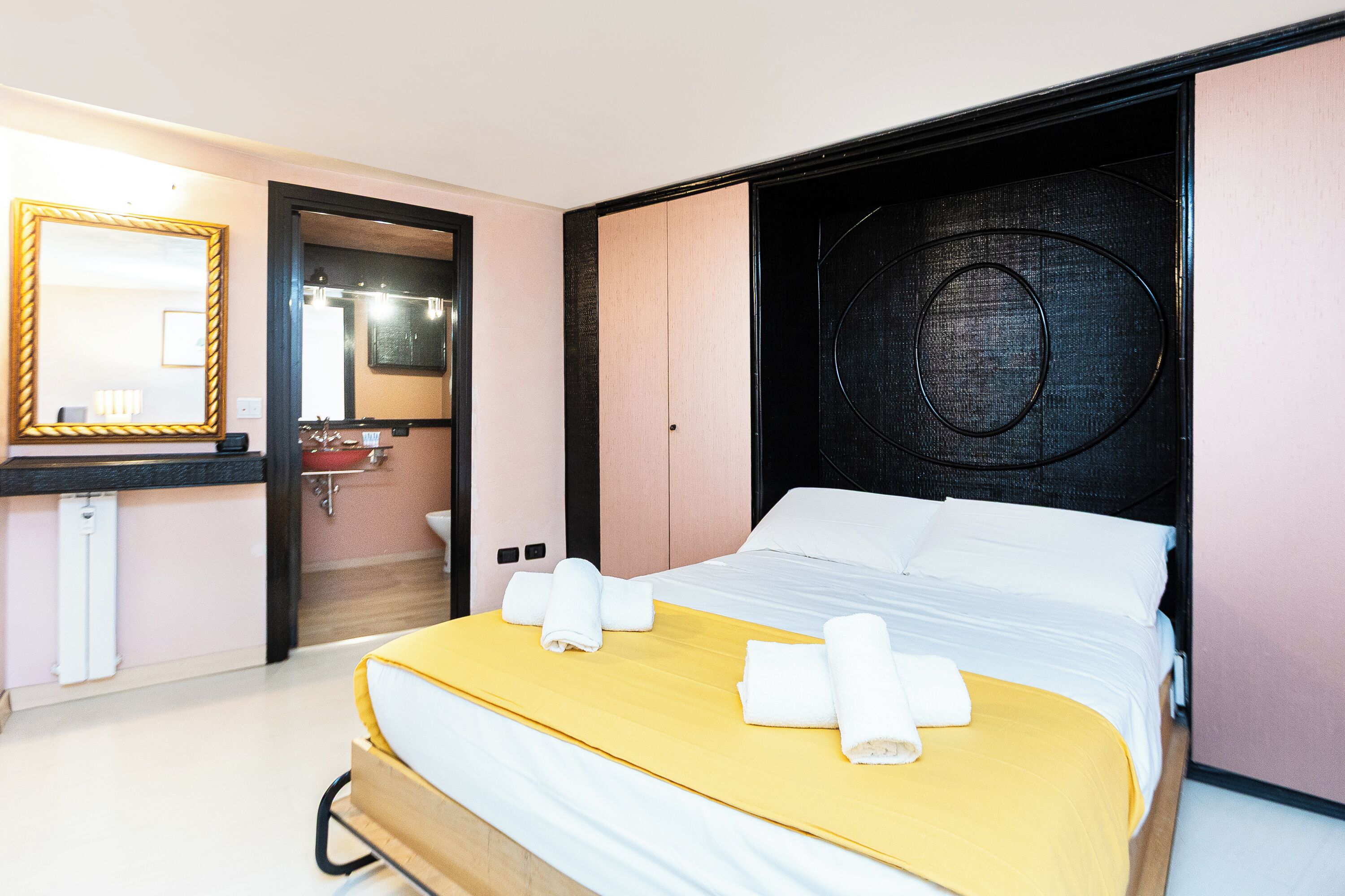 Quarto de hotel com cama, espelho, pia e porta que dá para outro cômodo.