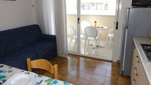 A compact living space with a sofa, dining table, chairs, a kitchen area with a refrigerator, and a sliding glass door leading to an outdoor balcony. - Great Flat 100mt From the sea (San Michele al Tagliamento)