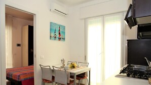 A compact hotel room with a kitchenette, a dining area with a table and chairs, a bed with a colorful bedspread, and a painting on the wall.