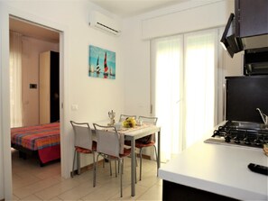 A compact hotel room with a kitchenette, a dining area with a table and chairs, a bed with a colorful bedspread, and a painting on the wall.