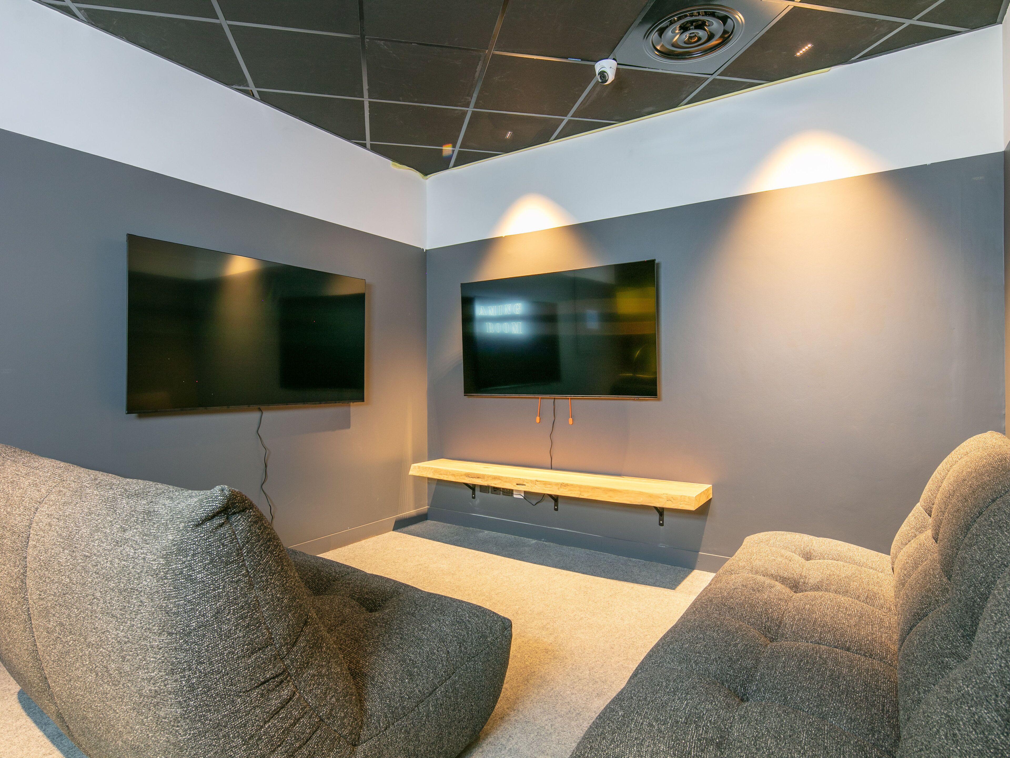 Ein modernes Zimmer mit zwei großen Flachbildfernsehern an der Wand, einem Holzregal darunter und zwei grauen Sitzsäcken.