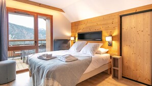 A bedroom with a bed, a TV mounted on the wall, a balcony with a view of the mountains, and a wooden floor. - T4 PMR | Skis aux pieds & espace bien-être (Vaujany)