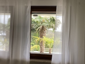 Una ventana con cortinas blancas traslúcidas, vista a una palmera y una planta pequeña en maceta.