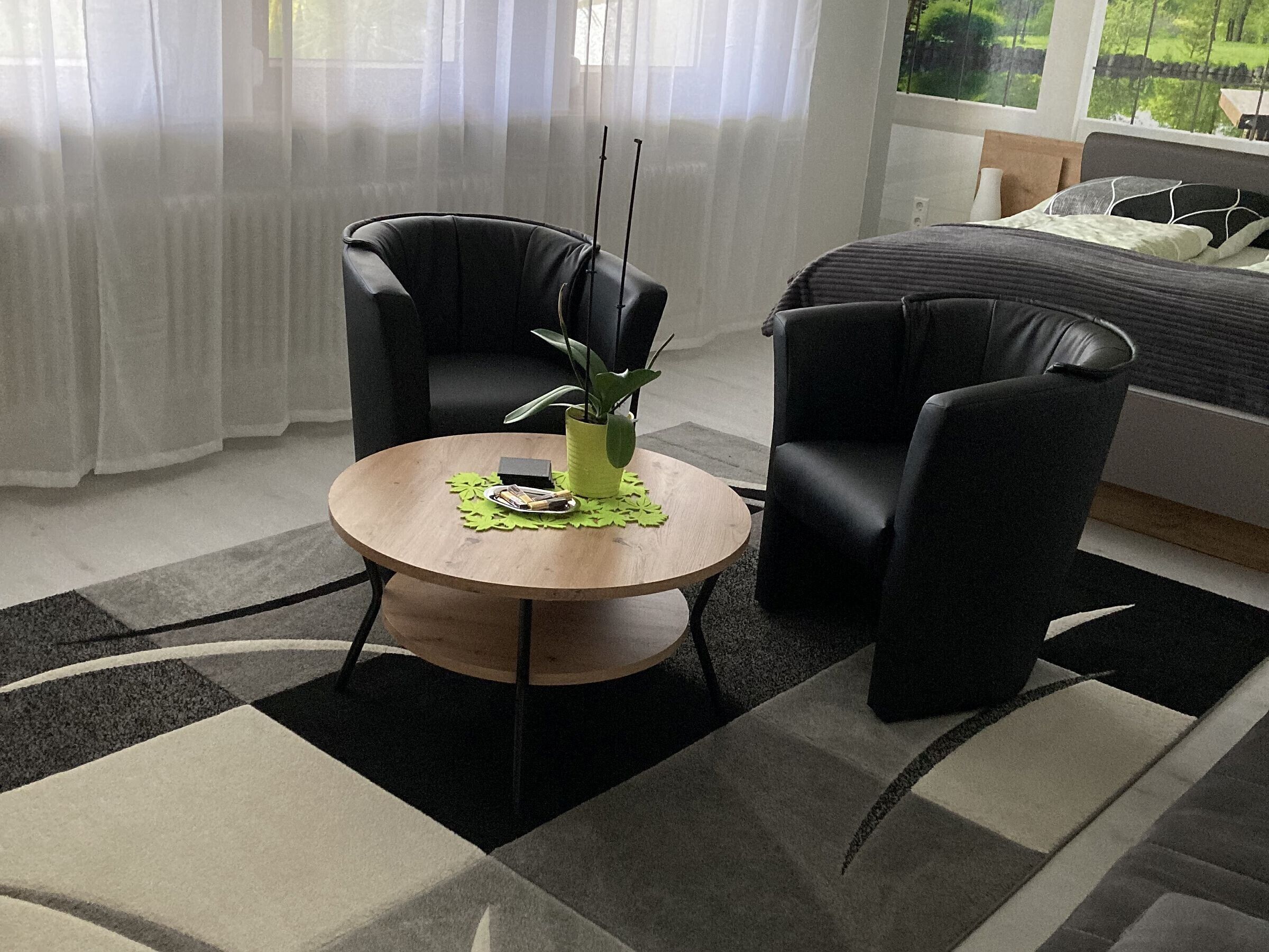 A modern hotel room with a round wooden table, two black leather chairs, a potted plant, and a bed with a grey headboard.