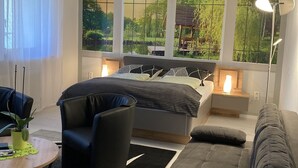 Een moderne slaapkamer met een groot bed, een bank, een stoel en een bureau. Er zijn ramen met uitzicht op groen en een houten constructie.