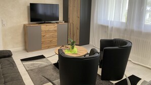Een moderne woonkamer met een flatscreen televisie, een houten kast, een zwarte bank, een ronde salontafel en een zwarte fauteuil.