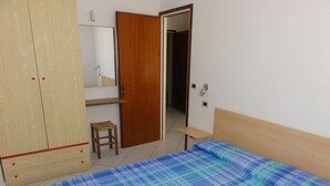 A bedroom with a bed, a wooden wardrobe, a small stool, a mirror, and a door leading to another room. - Sea View Flat Near Piazzale Zenith (San Michele al Tagliamento)