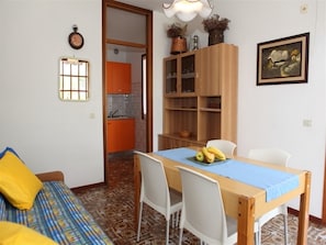 Un coin repas avec une table en bois, des chaises blanches et une nappe bleue. On aperçoit une cuisine à l’arrière-plan, avec des meubles de couleur orange.