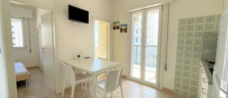 A compact kitchen with a white table and chairs, a flat-screen TV mounted above the counter, and a sliding glass door leading to a balcony.