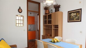 A dining area with a wooden table, white chairs, and a blue tablecloth. There is a kitchen in the background with orange cabinets.