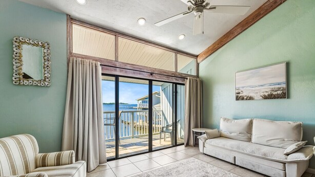 A spacious living room with a large sliding glass door leading to a balcony with a view of the water. There is a white sofa and striped armchairs, a small table, and a ceiling fan. - Top Floor 🪸 Private Balcony 🪸 View of the Gulf (Cedar Key)