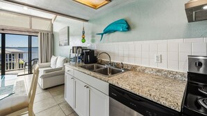 A modern kitchen with a granite countertop, stainless steel sink, and a dishwasher. There is a dining area with a white sofa and a view of the sea through large windows. - Top Floor 🪸 Private Balcony 🪸 View of the Gulf (Cedar Key)
