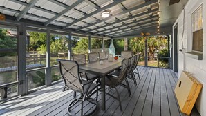 A covered outdoor dining area with a table and chairs, large windows, and a view of the surrounding greenery. - Cedar Key Seaside Escape (Cedar Key)