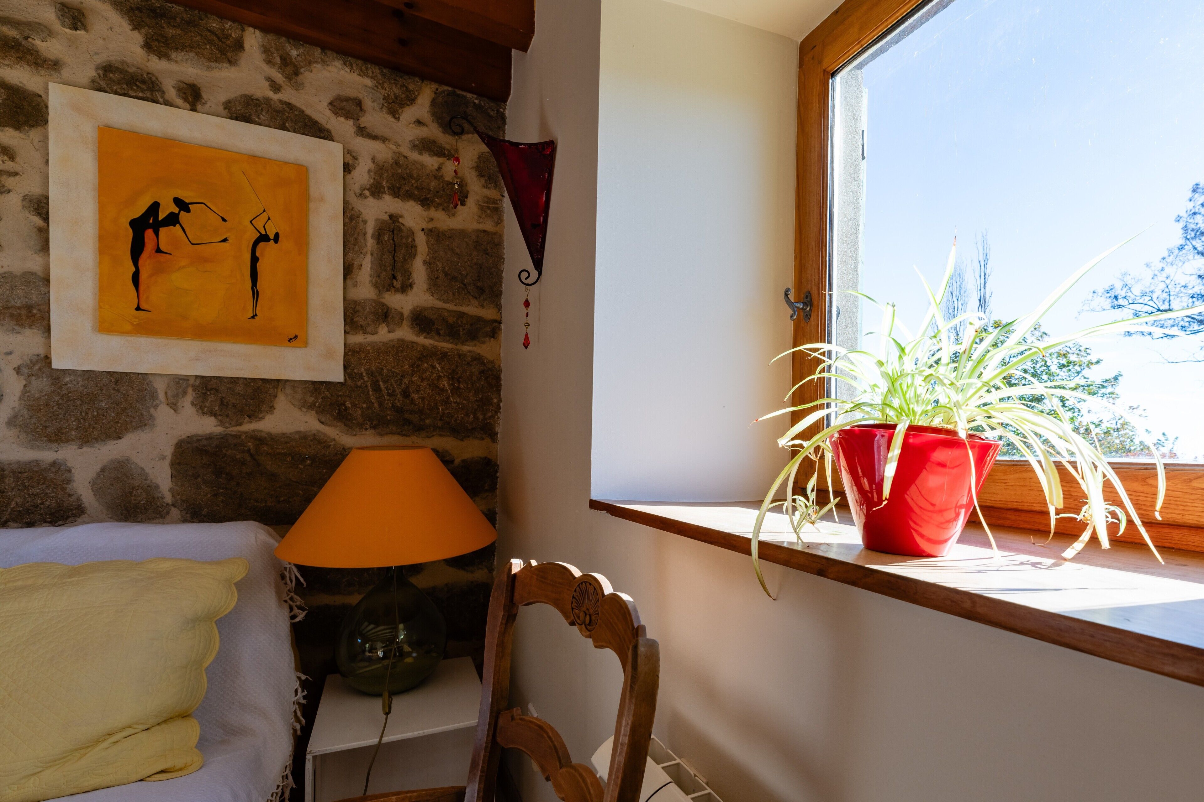 A room with a stone wall, a window with plants, a lamp, and a framed artwork.