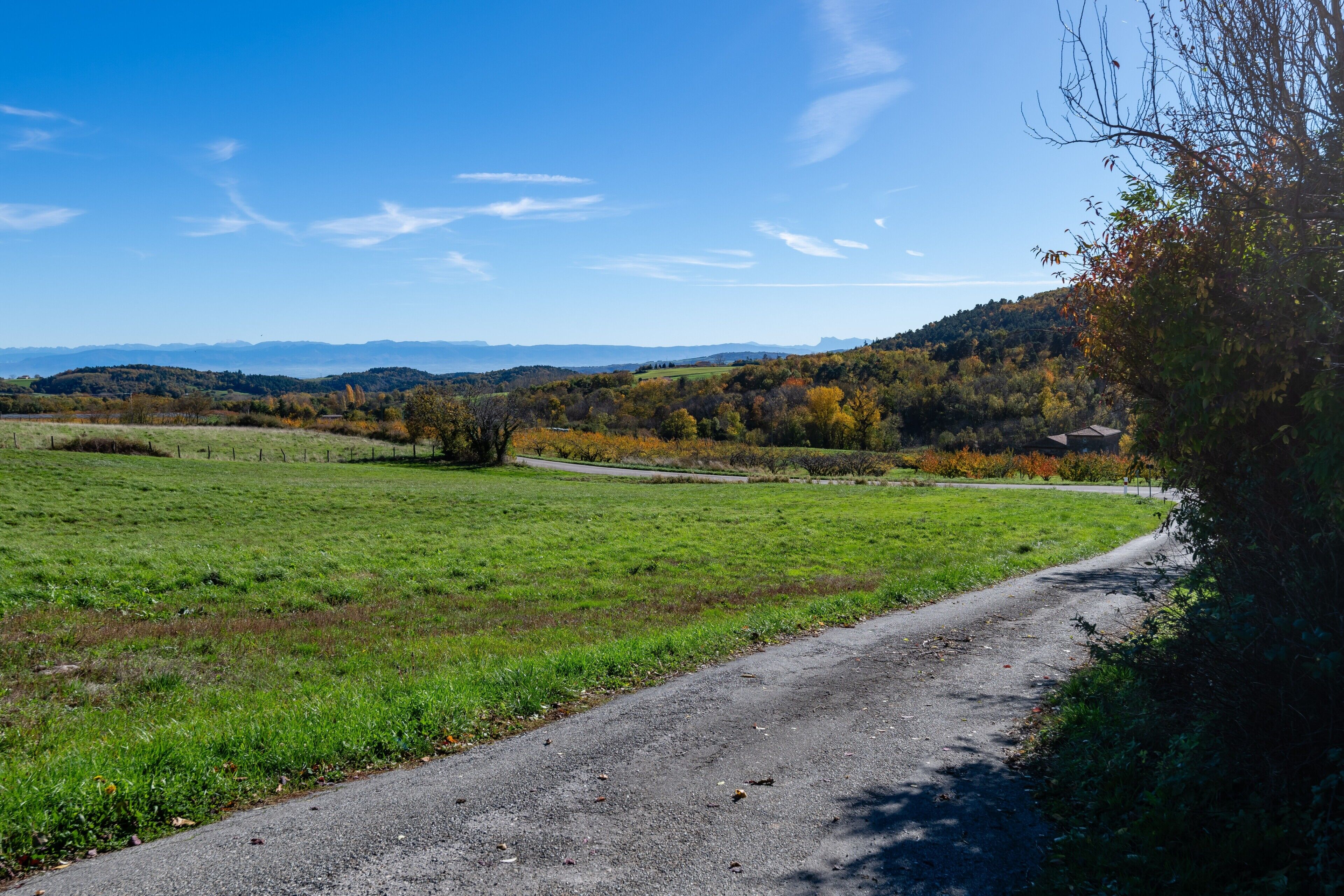 A rural road curving through a green field with distant hills and a clear sky.