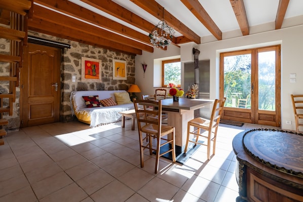 A cozy room with a wooden ceiling, stone walls, a bed with colorful pillows, a dining table with chairs, and a large window with a view of greenery.
