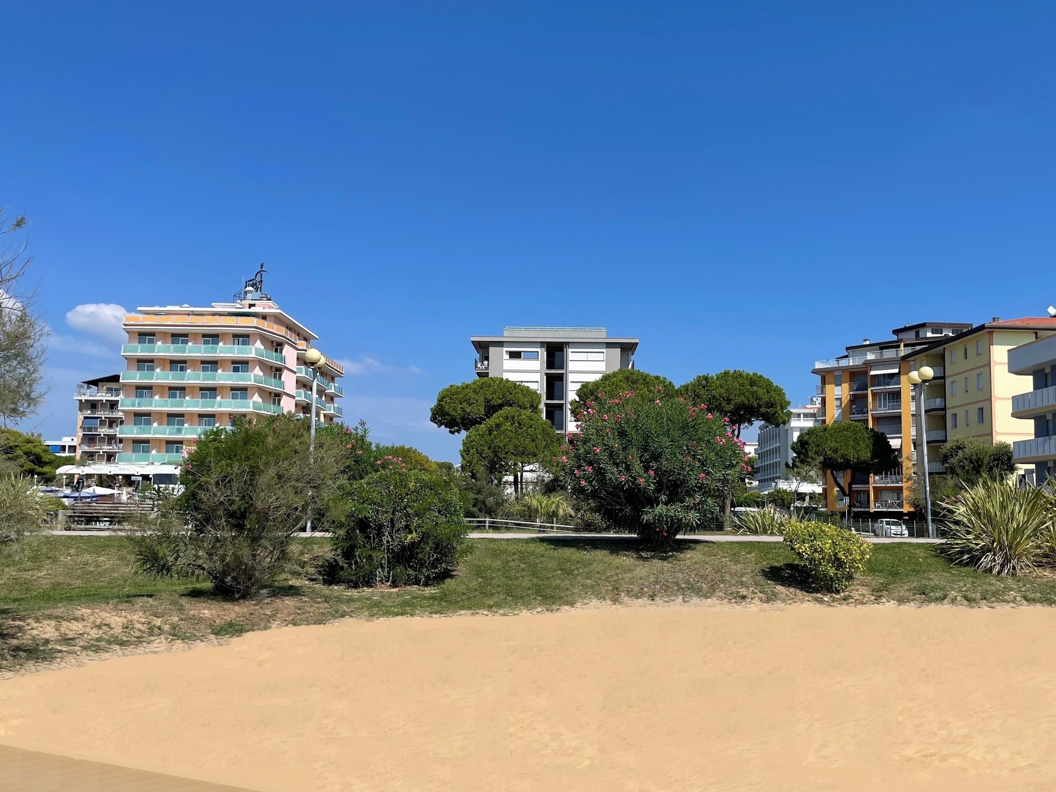 A coastal building with multiple balconies and a flat roof, surrounded by greenery and a sandy foreground.