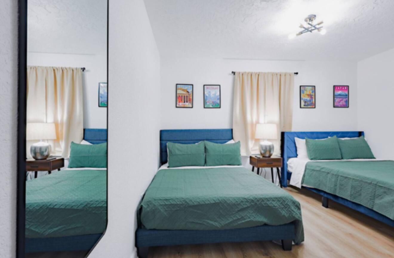 Une chambre avec deux lits simples, chacun doté d’une tête de lit bleue et de draps verts. On y trouve deux tables de chevet avec des lampes, et les murs sont décorés de tableaux encadrés.