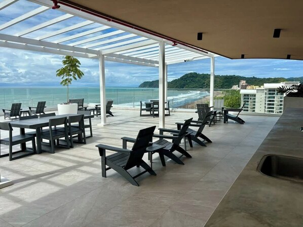 A covered outdoor seating area with tables and chairs, overlooking a beach and ocean.