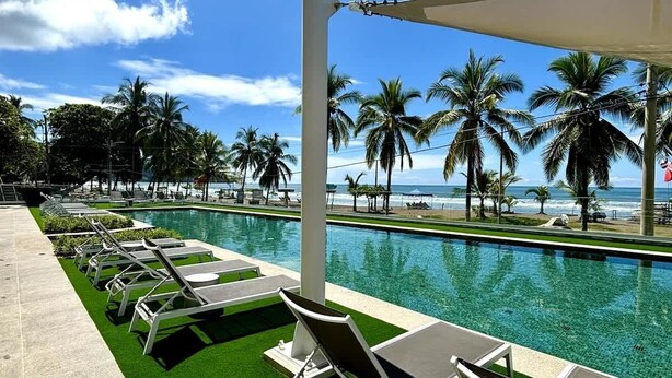 A poolside area with lounge chairs, a clear view of the ocean, and palm trees. - Stunning Beachfront 1BR Suite with Ocean View (Jacó)