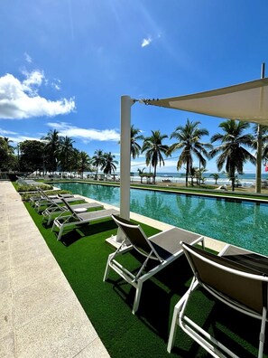 A poolside area with lounge chairs, a clear view of the ocean, and palm trees.