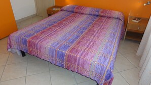 Een bed met een kleurrijk, patroonrijk beddegoed en kussens, een houten nachtkastje met een boek, en een deur links.