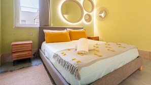 Ein ordentlich gemachtes Bett mit Decke und Kissen, ein Nachttisch, ein an der Wand befestigter Spiegel und ein Fenster mit Vorhängen.