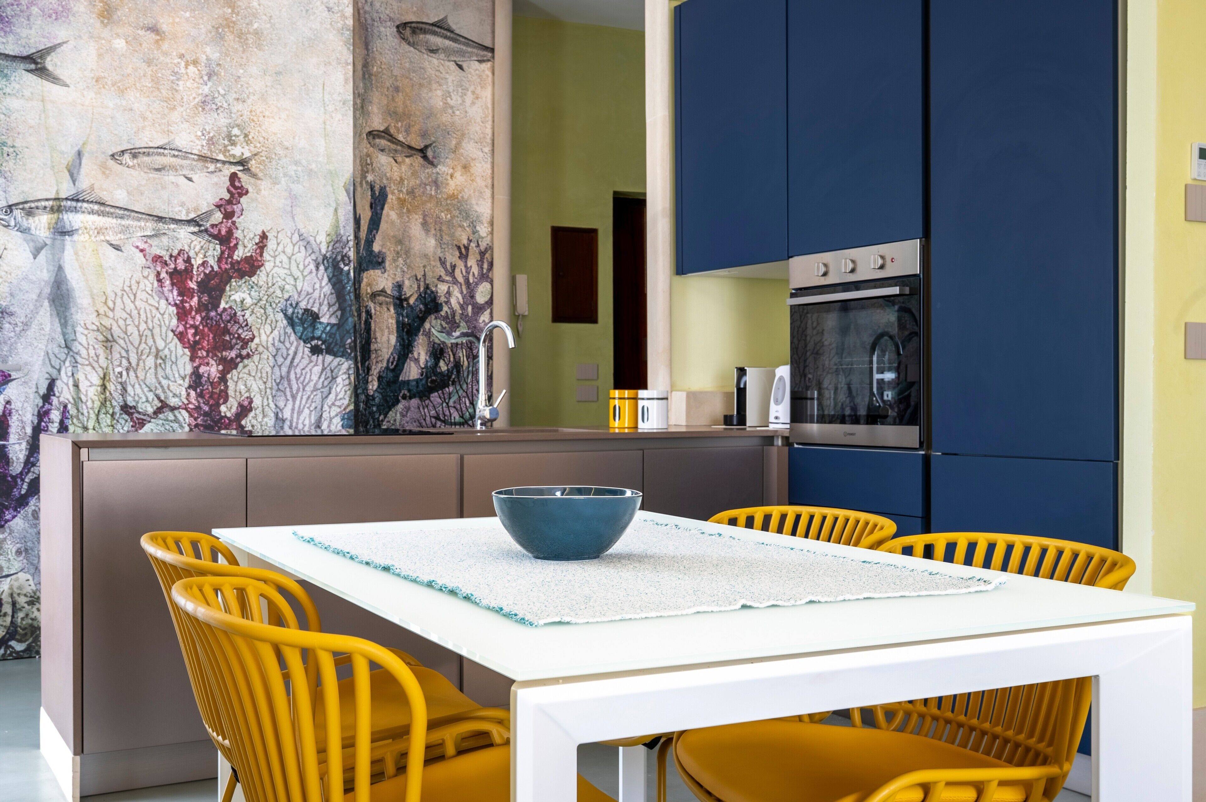 Une cuisine moderne avec une table blanche et des chaises jaunes, une grande fresque représentant des poissons et un four encastré.