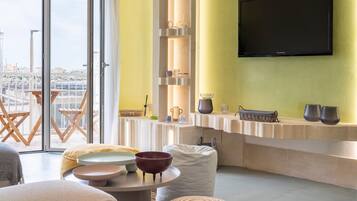 Un salon moderne doté d’un téléviseur, d’étagères ornées d’objets décoratifs, d’un coin repas avec une table et des chaises, et offrant une vue dégagée grâce à de grandes fenêtres.