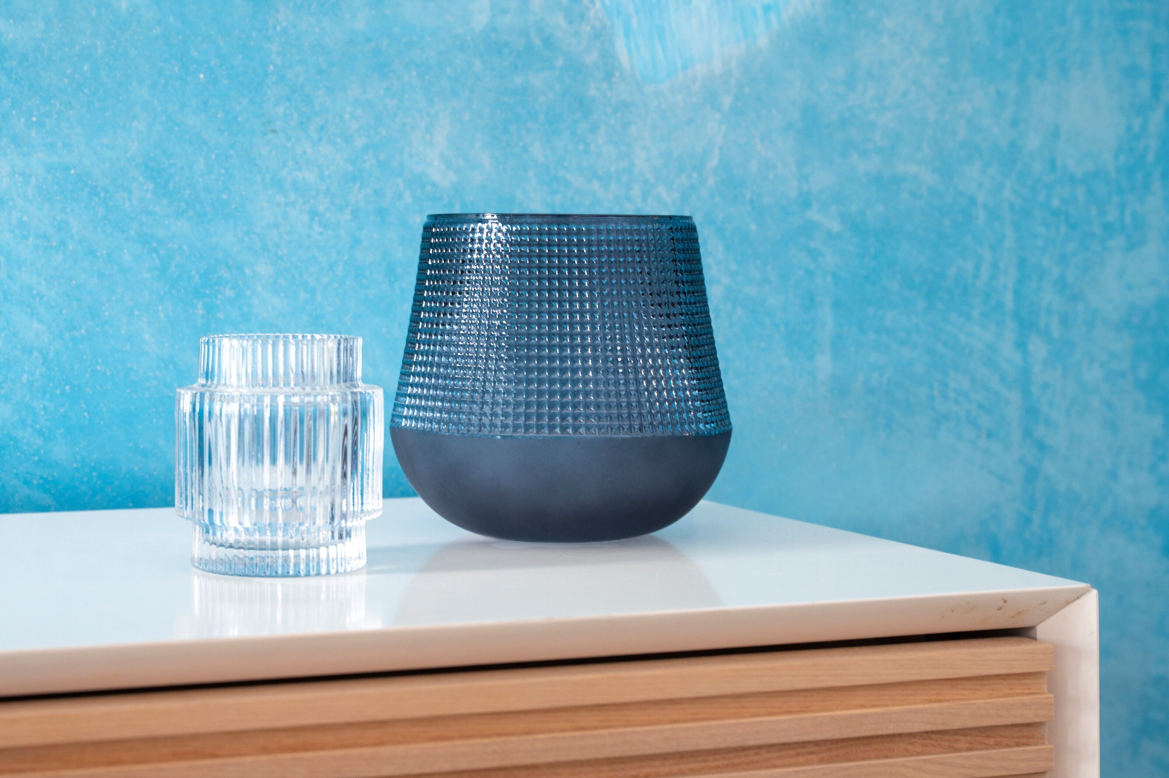 Un vaso transparente y un tazón azul con textura sobre una superficie blanca frente a una pared azul texturizada.
