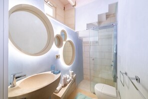 Et badeværelse med en stor, oval håndvask, flere runde spejle, en glasbrusekabine og et toilet.