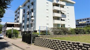 A multi-story residential building with balconies, a paved sidewalk, and a stone wall.