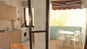 A compact kitchen with a washing machine, a dining area with a table and chairs, and a balcony with a view of trees.
