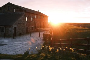A stone building with a tiled entrance, a wooden fence, and a sunrise in the background. - Cobbled Curlew, Patty's Barn , Lancaster 4* (Lancaster)