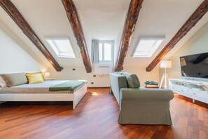 Une chambre aménagée en mezzanine comprenant un lit, un canapé et un téléviseur, avec des poutres apparentes et des fenêtres de toit.