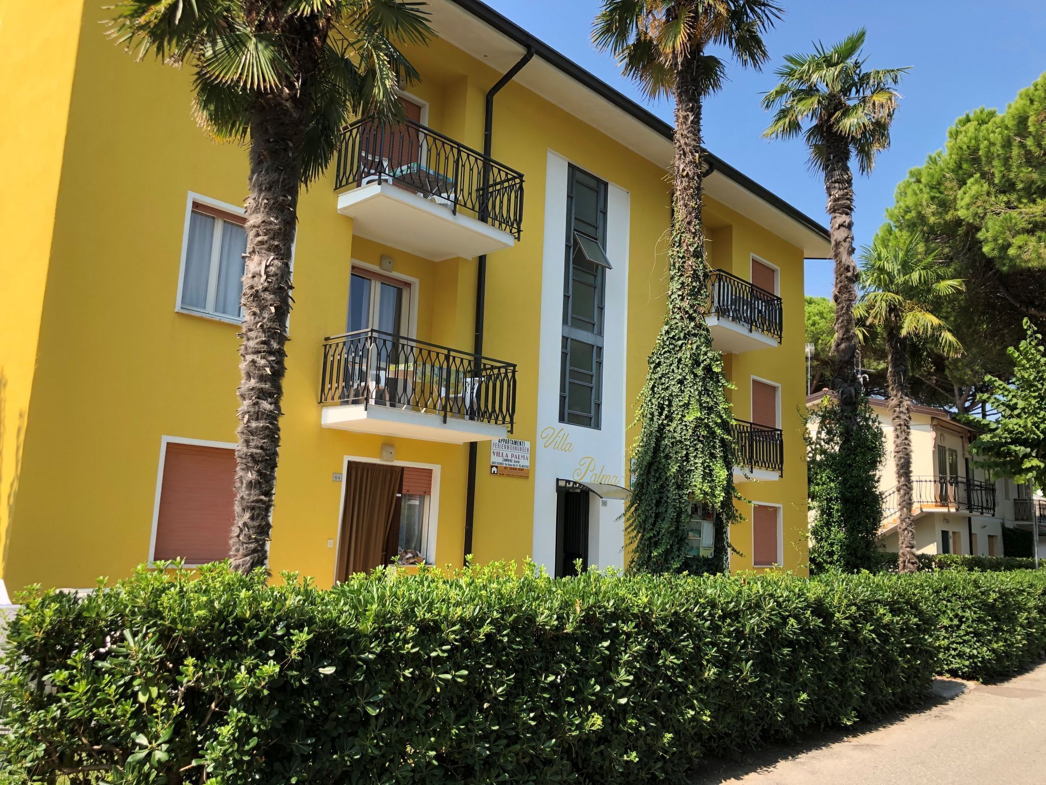 Un bâtiment jaune doté de balcons, une enseigne indiquant « Villa Palma », et une végétation luxuriante tout autour.