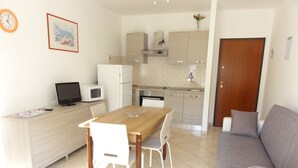 A compact living space with a kitchenette, a dining area with a wooden table and white chairs, and a grey sofa. - Bright Flat in a Quiet Area Near the Beach (San Michele al Tagliamento)