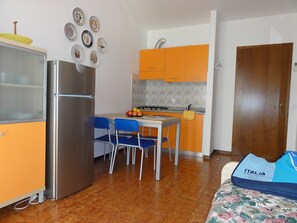 Eine kleine Küche mit orangefarbenen Schränken, einem Edelstahlkühlschrank und einem Essbereich mit blauen Stühlen.