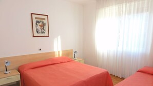 A hotel room with two beds, a nightstand with a lamp, a framed picture on the wall, and a window with sheer curtains.