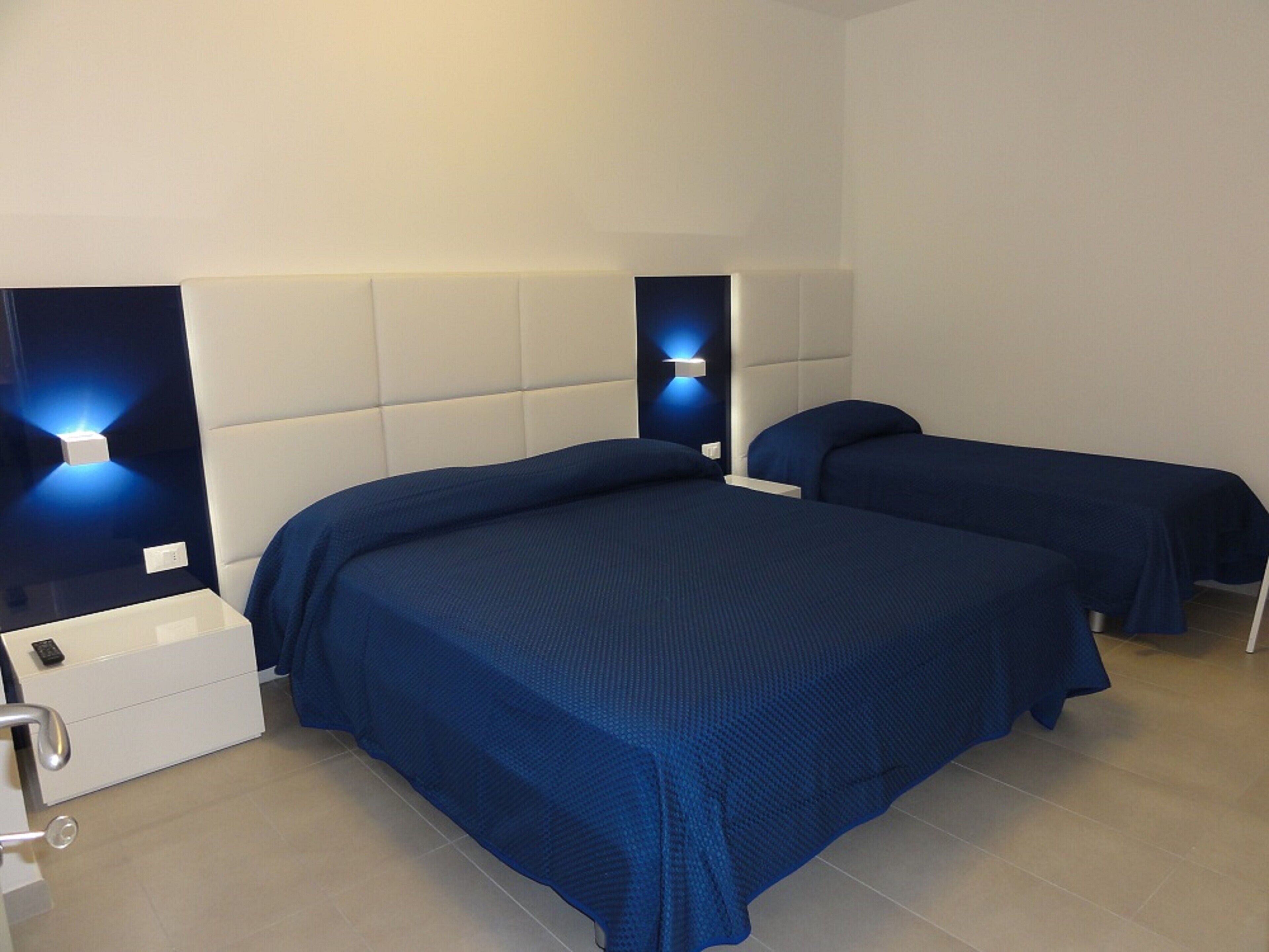 A hotel room with two beds, a white chest of drawers, and a bedside table with a lamp.