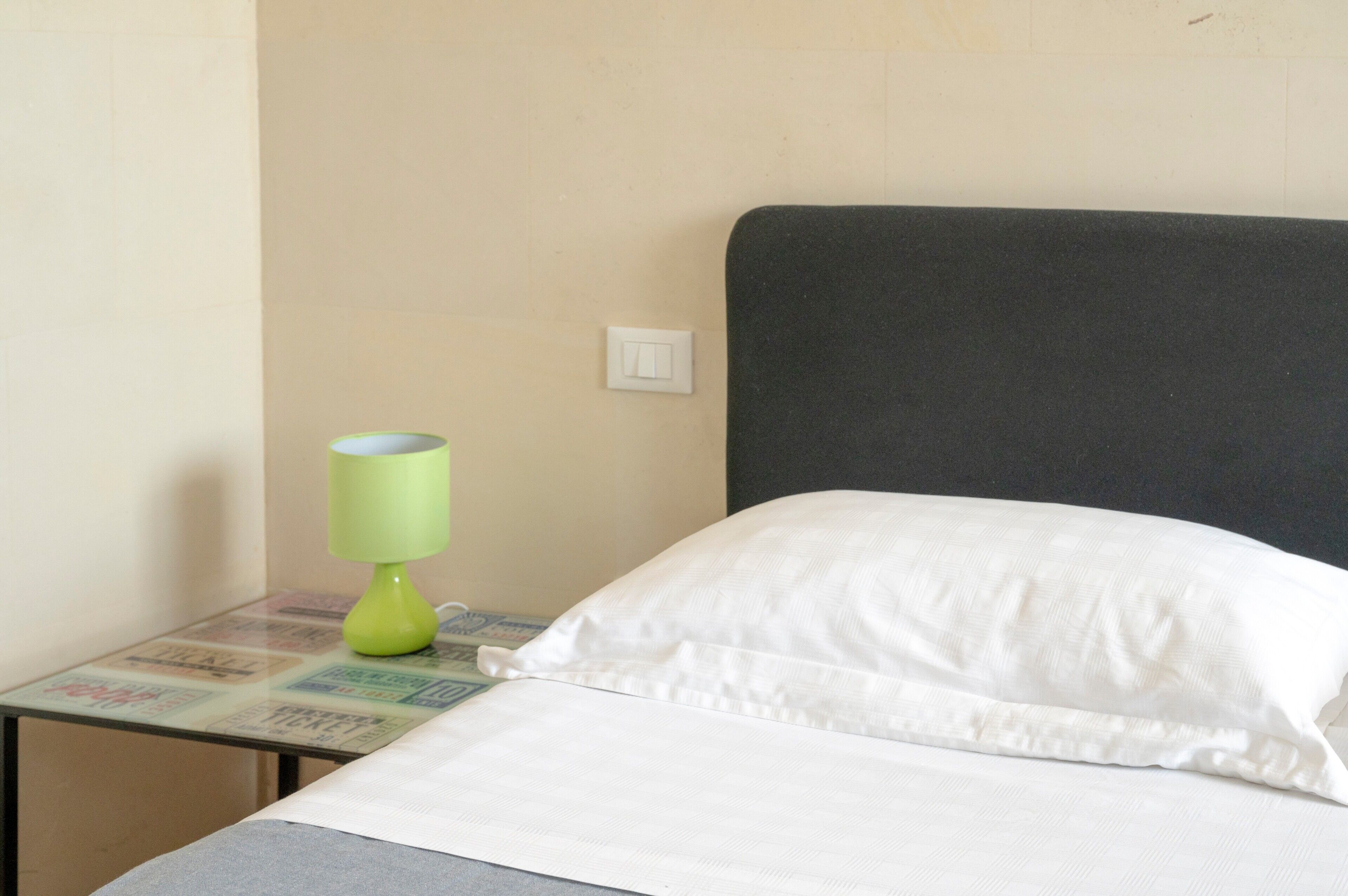 Ein Bett mit weißer Bettwäsche, eine grüne Lampe auf einem kleinen Nachttisch daneben und ein Lichtschalter an der Wand.