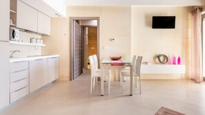 Una cocina moderna con armarios blancos, un área de comedor con mesa de vidrio y un televisor de pantalla plana montado en la pared.