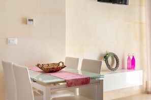 A dining area with a glass table, white chairs, a woven basket, a pink tablecloth, a wall-mounted TV, and a shelf with two bottles.