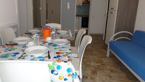 A dining area with a table set for four, chairs, a blue sofa, and a kitchen in the background. - Flat in a Modern Coastal Building (San Michele al Tagliamento)