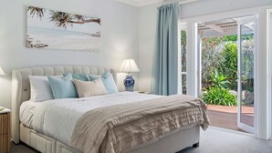 A bedroom with a bed, a nightstand, a lamp, a window with curtains, and a view of a garden.
