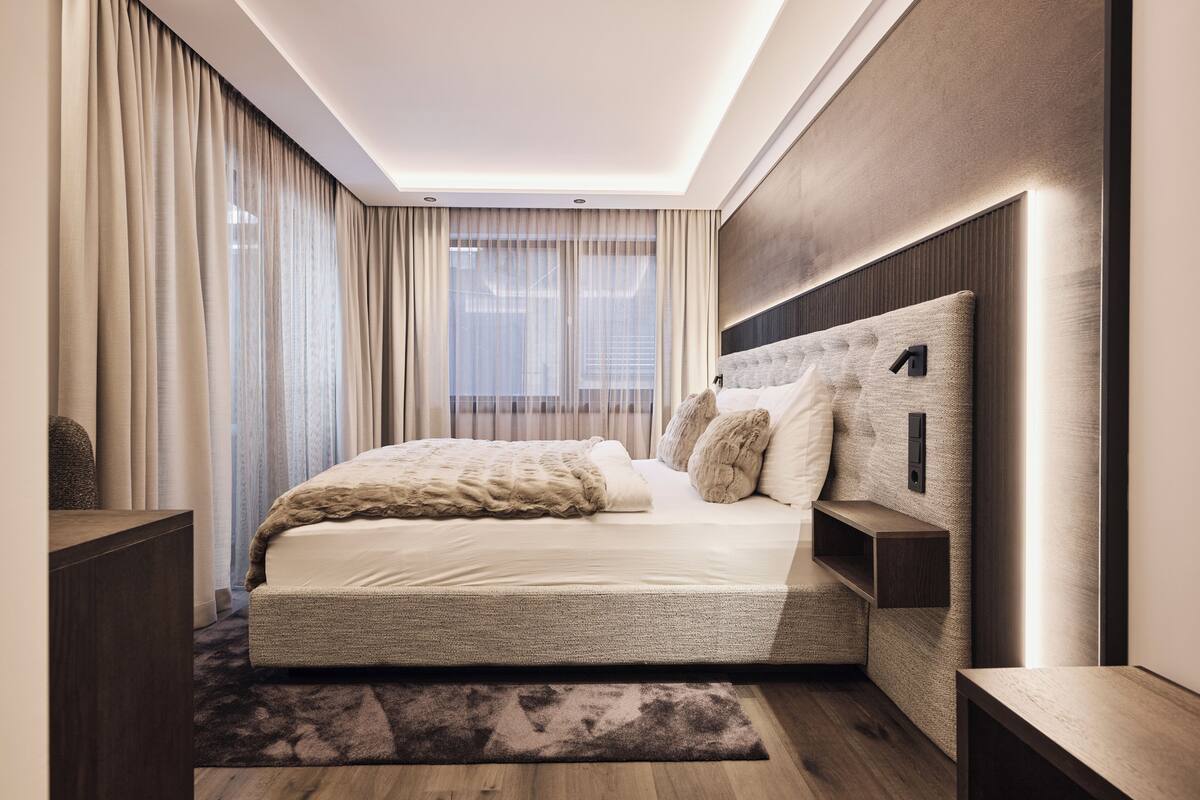 Ein modernes Schlafzimmer mit einem großen Bett, einer Kopfteil mit integrierter Beleuchtung, einem Nachttisch und Blick ins Freie durch transparente Vorhänge.