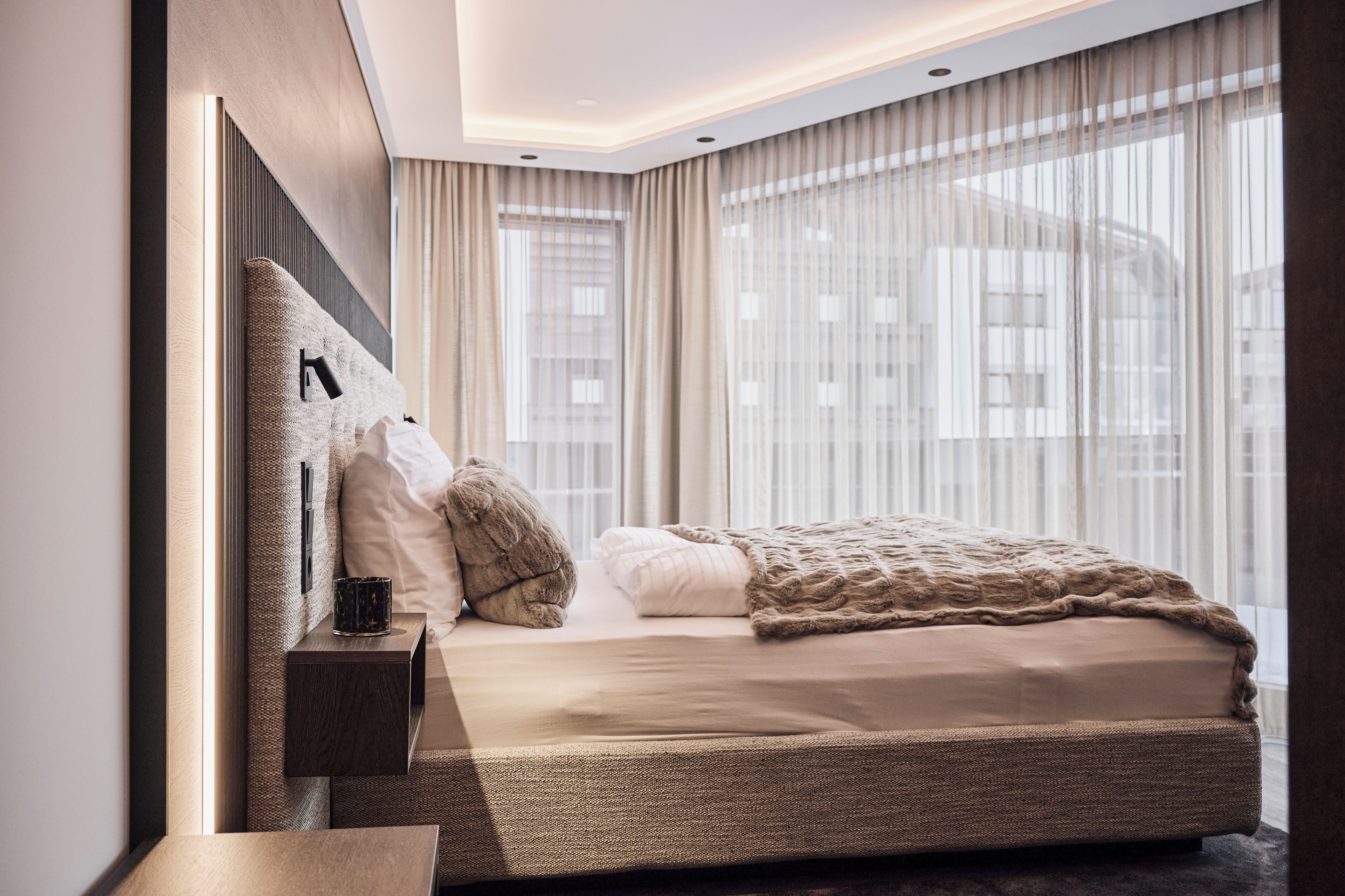 Une chambre moderne avec un grand lit, une table de chevet et une vue sur la ville à travers de fines rideaux.