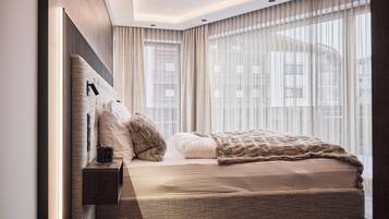 Une chambre moderne avec un grand lit, une table de chevet et une vue sur la ville à travers de fines rideaux.