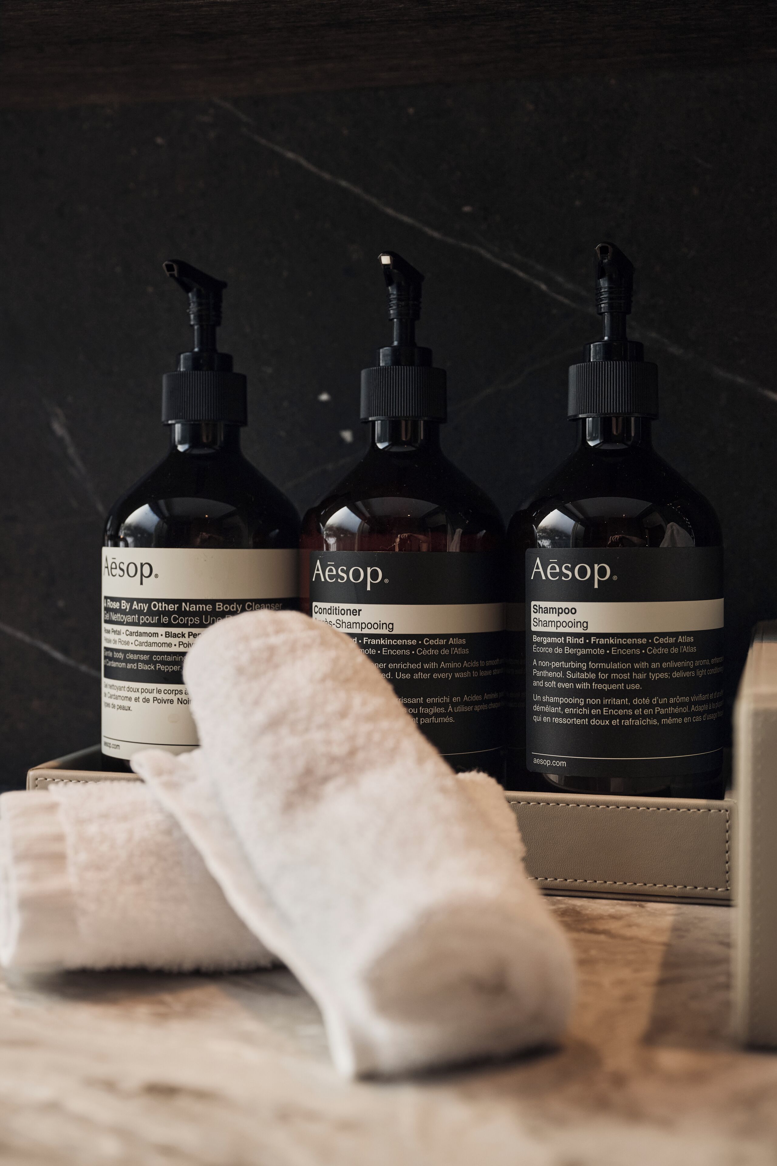 Drei Flaschen Aesop Körperpflegeprodukte und ein aufgerolltes Handtuch auf einer dunklen Oberfläche.